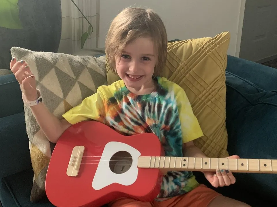 A smiling young child playing a red 3-string guitar sized for small hands